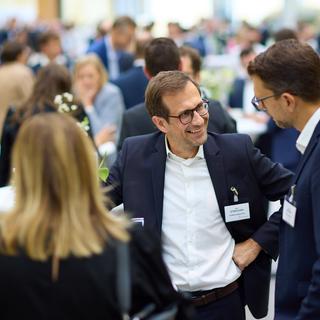 Austrian CFO Day 2025 Drei Personen in Businesskleidung stehen in einem belebten Raum mit vielen weiteren Menschen im Hintergrund, die an Stehtischen stehen und sich unterhalten.