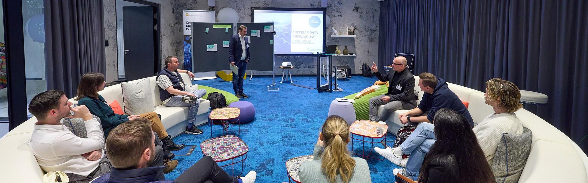 Austrian Data_Business_Day 2025 Mehrere Personen sitzen in einem großen, runden weißen Sofa in einem modernen Raum mit blauem Teppich, während ein Mann vor einer Präsentation steht.