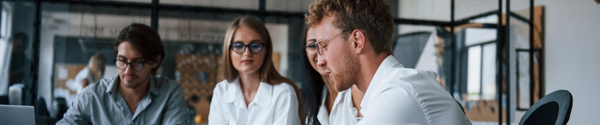 Drei Personen sitzen an einem Tisch in einem modernen Büro und schauen auf einen Laptop Drei Personen sitzen an einem Tisch in einem modernen Büro und schauen auf einen Laptop