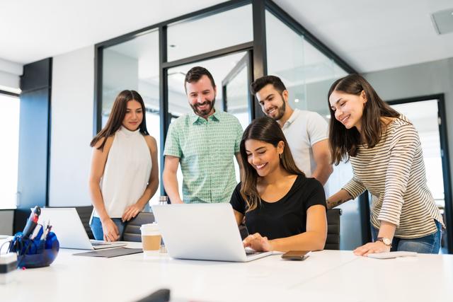 Gruppe von vier Personen, zwei Frauen sitzen, zwei Männer stehen, alle blicken auf einen Laptop auf einem Tisch in einem modernen Büro Gruppe von vier Personen, zwei Frauen sitzen, zwei Männer stehen, alle blicken auf einen Laptop auf einem Tisch in einem modernen Büro
