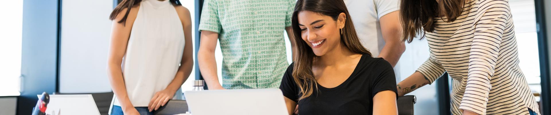 Gruppe von vier Personen, zwei Frauen sitzen, zwei Männer stehen, alle blicken auf einen Laptop auf einem Tisch in einem modernen Büro Gruppe von vier Personen, zwei Frauen sitzen, zwei Männer stehen, alle blicken auf einen Laptop auf einem Tisch in einem modernen Büro
