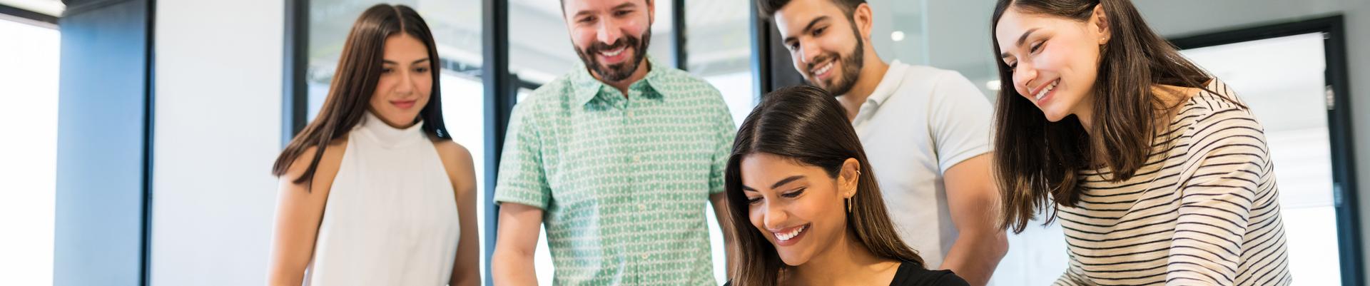 Gruppe von vier Personen, zwei Frauen sitzen, zwei Männer stehen, alle blicken auf einen Laptop auf einem Tisch in einem modernen Büro