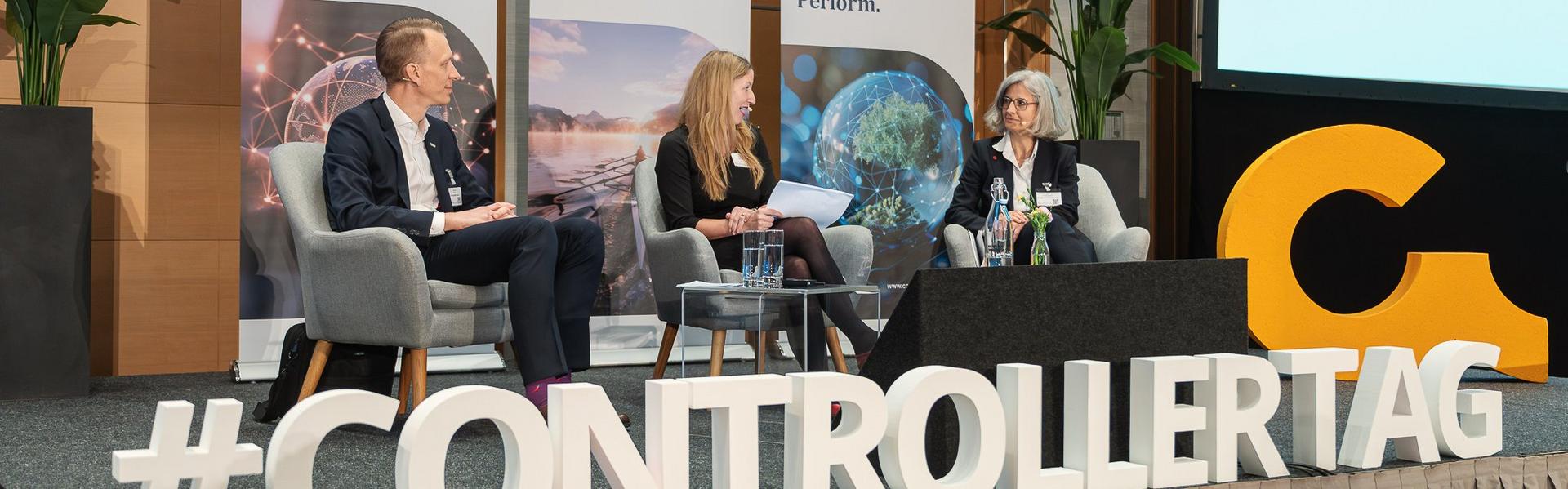 Controllertag_2026 Drei Personen sitzen auf Sesseln auf einer Bühne vor Bannern mit dem Text 'Connect. Engage. Perform.' und großen Buchstaben '#CONTROLLER TAG' im Vordergrund