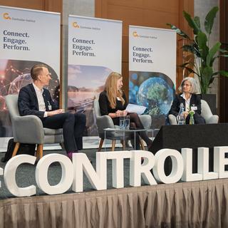 Drei Personen sitzen auf Sesseln auf einer Bühne vor Bannern mit dem Text 'Connect. Engage. Perform.' und großen Buchstaben '#CONTROLLER TAG' im Vordergrund