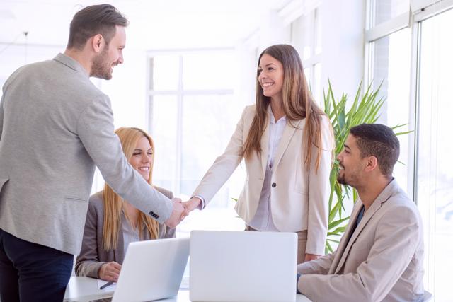 Vier Personen in einem Bürogebäude, zwei stehen und geben sich die Hand, zwei sitzen am Tisch und beobachten Vier Personen in einem Bürogebäude, zwei stehen und geben sich die Hand, zwei sitzen am Tisch und beobachten