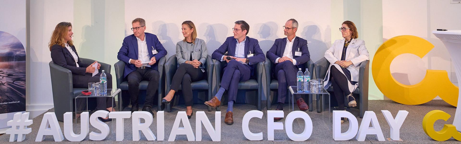 Austrian CFO Day 2025 Sechs Personen sitzen auf einer Bühne bei einer Podiumsdiskussion mit dem Schriftzug '#AUSTRIAN CFO DAY' vorne auf der Bühne und zwei Bannern mit dem Text 'Connect. Engage. Perform.'
