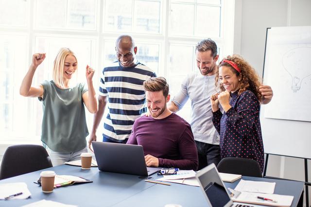 Fünf Personen in einem Büro feiern gemeinsam vor einem Laptop, sichtbar erfreut und motiviert