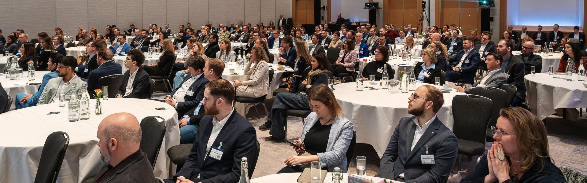 Controllertag_2026 Großer Konferenzraum mit mehreren runden Tischen, an denen zahlreiche Personen in Businesskleidung sitzen und einem Vortrag folgen