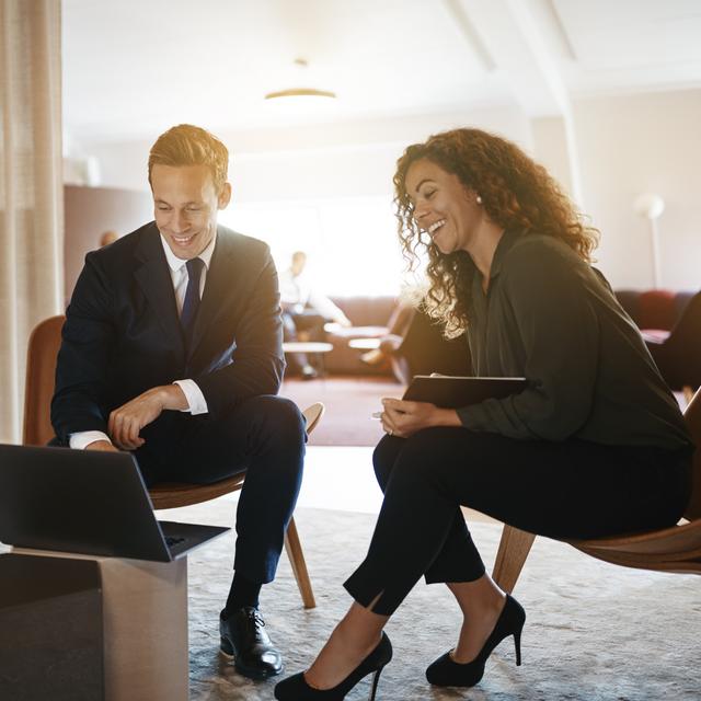 Zwei Geschäftsleute in formeller Kleidung sitzen auf Stühlen und schauen lächelnd auf einen Laptop in einem hellen Büro