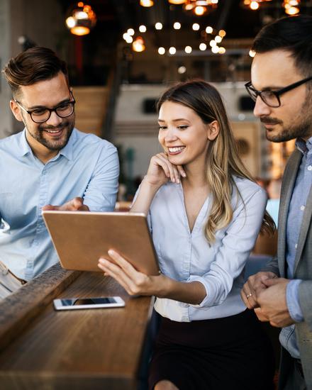 Drei junge Berufstätige in Büro mit Tablet, eine Frau zeigt zwei Männern etwas auf dem Bildschirm, alle lächeln