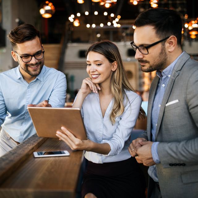 Drei junge Berufstätige in Büro mit Tablet, eine Frau zeigt zwei Männern etwas auf dem Bildschirm, alle lächeln