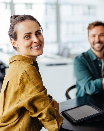 Drei lächelnde Personen sitzen an einem Tisch in einem modernen Büro, eine Frau im Vordergrund blickt zur Kamera