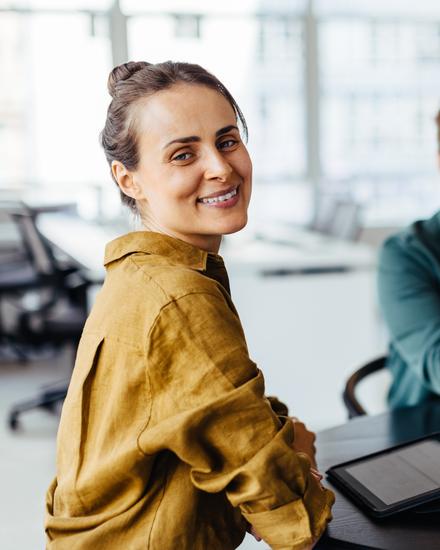 Drei lächelnde Personen sitzen an einem Tisch in einem modernen Büro, eine Frau im Vordergrund blickt zur Kamera