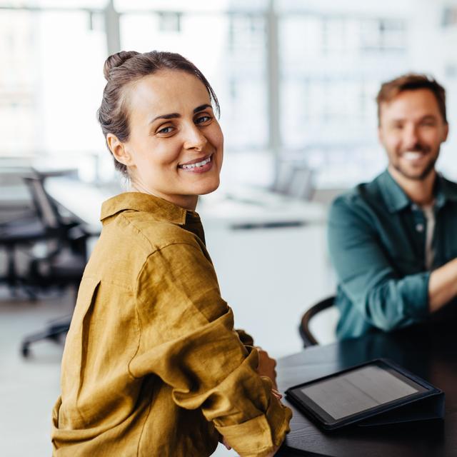 Drei lächelnde Personen sitzen an einem Tisch in einem modernen Büro, eine Frau im Vordergrund blickt zur Kamera