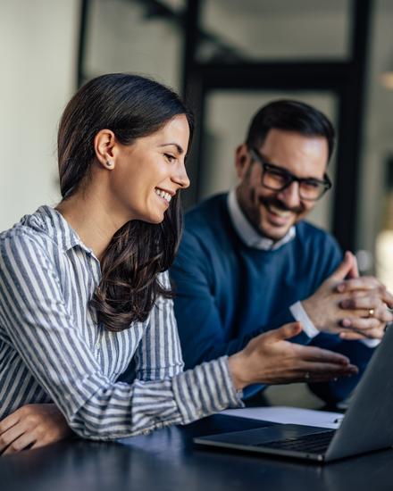 Zwei lächelnde Personen, eine Frau und ein Mann, arbeiten gemeinsam an einem Laptop in einem modernen Büro