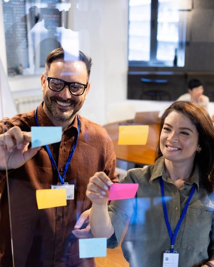 Zwei Personen lächeln und zeigen auf Post its an einer Glaswand in einem modernen Büro mit Sitzgelegenheiten im Hintergrund
