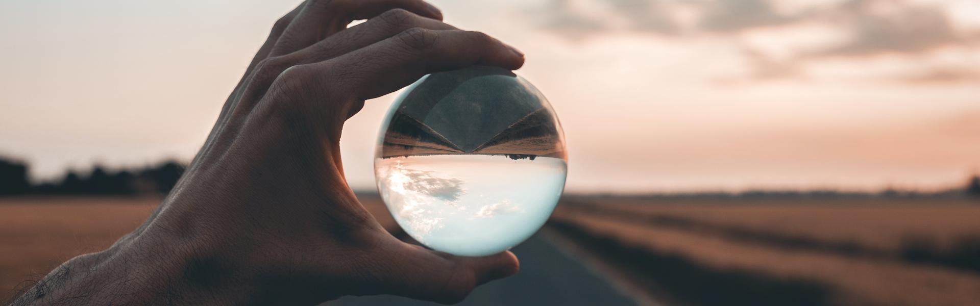 Sujet_Austrian_CFO_Day_2025 Eine Hand hält eine Kristallkugel, die eine Landschaft mit einem Weg und einem Himmel mit Wolken reflektiert.