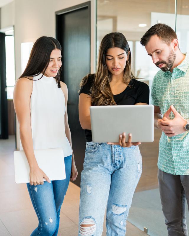 Drei junge Erwachsene stehen in einem hellen Raum und schauen gemeinsam auf einen Laptop