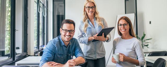 Drei Personen sitzen an einem Tisch in einem modernen Büro mit großen Fenstern und arbeiten gemeinsam an einem Laptop Drei Personen sitzen an einem Tisch in einem modernen Büro mit großen Fenstern und arbeiten gemeinsam an einem Laptop