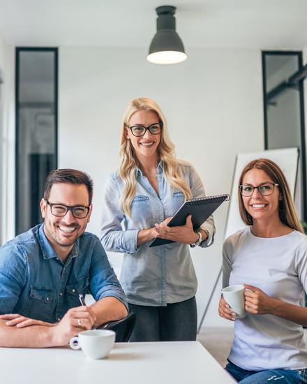 Drei Personen sitzen an einem Tisch in einem modernen Büro mit großen Fenstern und arbeiten gemeinsam an einem Laptop