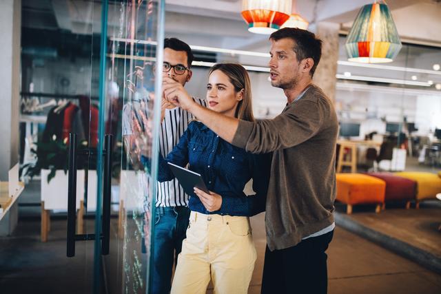 Drei junge Erwachsene diskutieren und zeigen auf eine Glaswand mit Notizen in einem modernen Büro