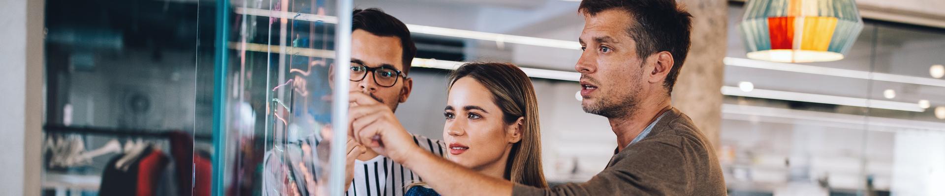 Drei junge Erwachsene diskutieren und zeigen auf eine Glaswand mit Notizen in einem modernen Büro