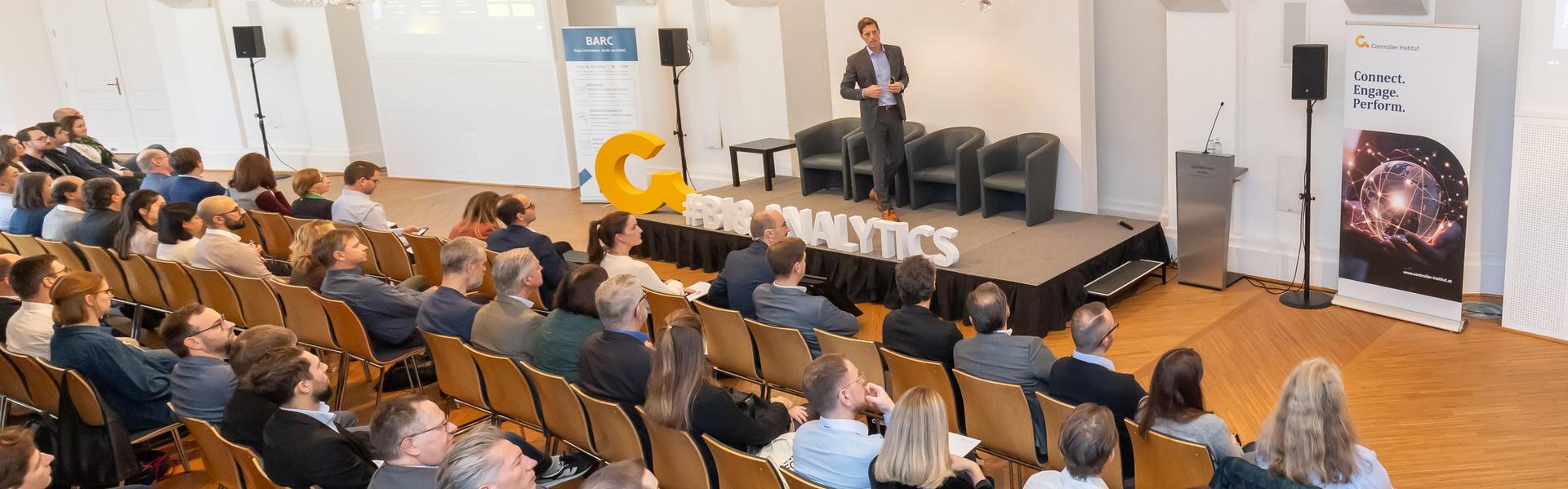 Mann steht auf Bühne vor Publikum in hellem Saal mit Kronleuchtern, Banner mit Netzwerk- und Technologie-Motiv rechts.