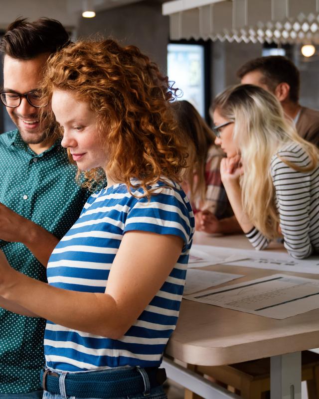 Zwei junge Erwachsene, die gemeinsam auf ein Tablet schauen, während zwei weitere Personen im Hintergrund an einem Stehtisch arbeiten
