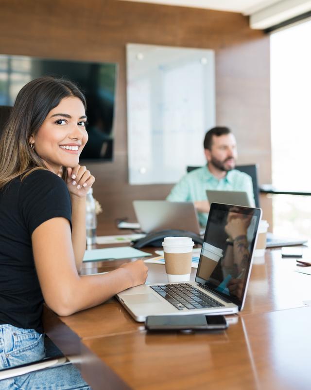 Frau mit schwarzem T-Shirt sitzt an einem Tisch mit Laptop und lächelt in einem modernen Büro mit mehreren Personen im Hintergrund Frau mit schwarzem T-Shirt sitzt an einem Tisch mit Laptop und lächelt in einem modernen Büro mit mehreren Personen im Hintergrund