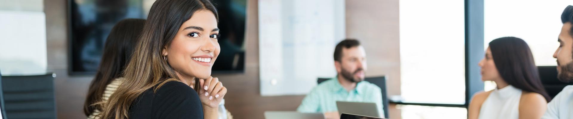 Frau mit schwarzem T-Shirt sitzt an einem Tisch mit Laptop und lächelt in einem modernen Büro mit mehreren Personen im Hintergrund Frau mit schwarzem T-Shirt sitzt an einem Tisch mit Laptop und lächelt in einem modernen Büro mit mehreren Personen im Hintergrund