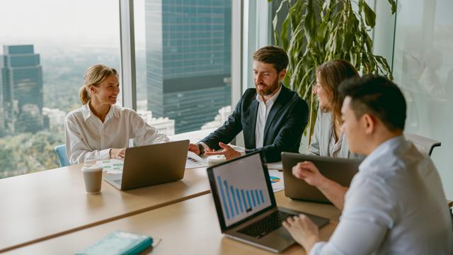 Drei Personen sitzen an einem Tisch, zwei mit Laptops, in einem modernen Büro mit großen Fenstern und Stadtblick Drei Personen sitzen an einem Tisch, zwei mit Laptops, in einem modernen Büro mit großen Fenstern und Stadtblick