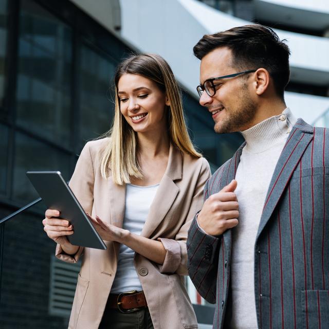 Zwei junge Berufstätige, eine Frau und ein Mann, sehen gemeinsam auf ein Tablet und lächeln, draußen vor modernen Gebäuden