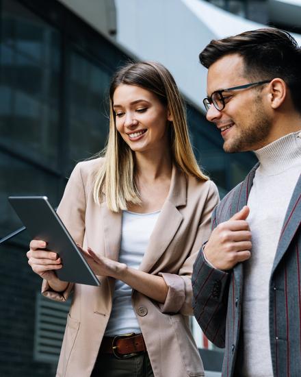 Zwei junge Berufstätige, eine Frau und ein Mann, sehen gemeinsam auf ein Tablet und lächeln, draußen vor modernen Gebäuden