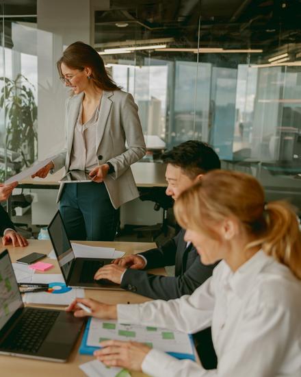 Drei Personen sitzen an einem Tisch mit Laptops und Unterlagen, eine steht und spricht zu den anderen in einem hellen Büro mit großen Fenstern