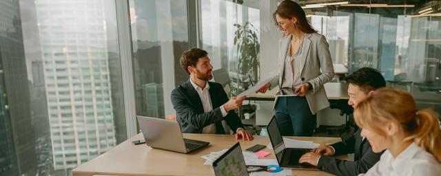 Drei Personen sitzen an einem Tisch mit Laptops und Unterlagen, eine steht und spricht zu den anderen in einem hellen Büro mit großen Fenstern