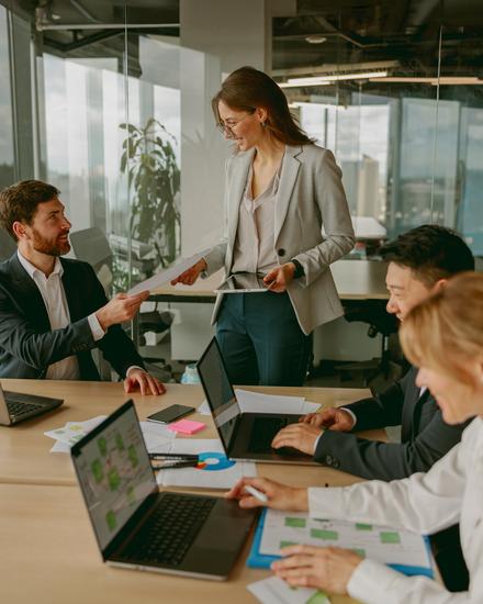 Drei Personen sitzen an einem Tisch mit Laptops und Unterlagen, eine steht und spricht zu den anderen in einem hellen Büro mit großen Fenstern