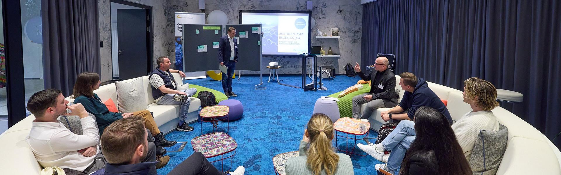 Mehrere Personen sitzen in einem großen, runden weißen Sofa in einem modernen Raum mit blauem Teppich, während ein Mann vor einer Präsentation steht.