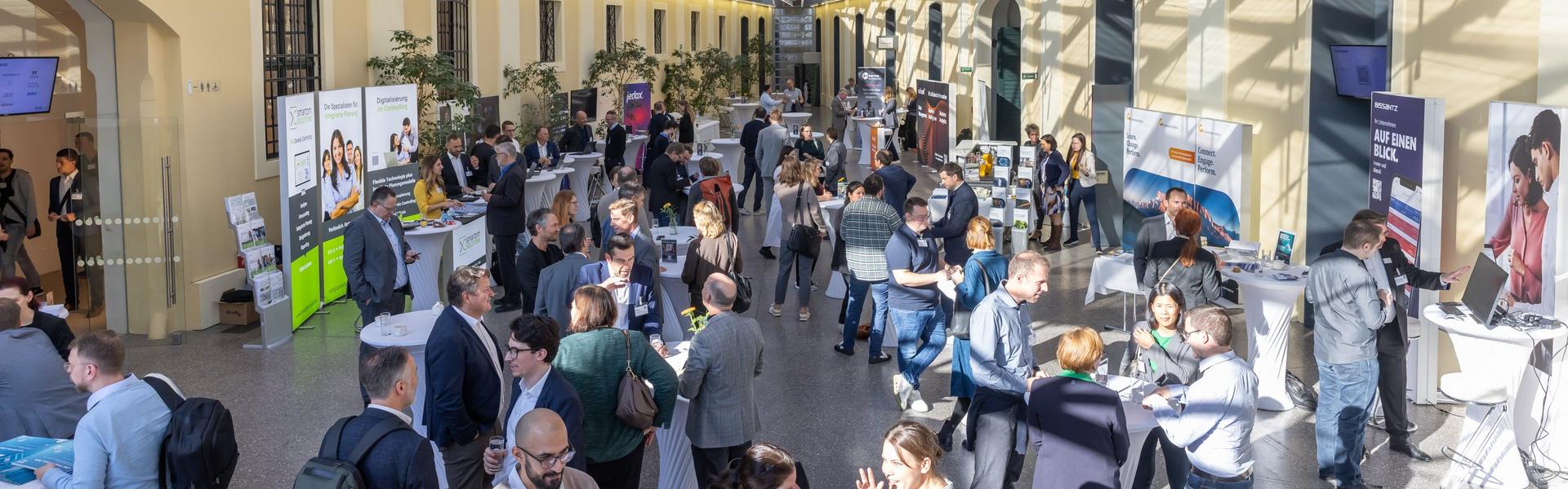 Großer, heller Innenraum mit Glasdach, vielen Menschen an Stehtischen und Informationsständen, gelbe Wände mit Rundbögen.
