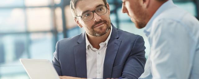 Zwei Männer im Gespräch, einer hält ein Tablet, beide tragen Hemden und sitzen an einem Tisch in einem hellen Büro Zwei Männer im Gespräch, einer hält ein Tablet, beide tragen Hemden und sitzen an einem Tisch in einem hellen Büro