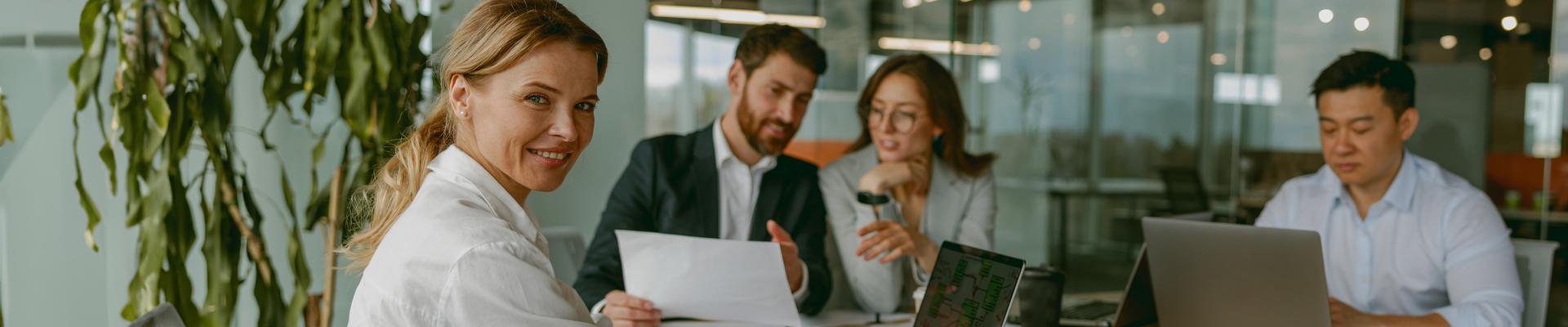 Drei Personen sitzen an einem Tisch in einem Büro, eine Person schaut zur Kamera und lächelt Drei Personen sitzen an einem Tisch in einem Büro, eine Person schaut zur Kamera und lächelt