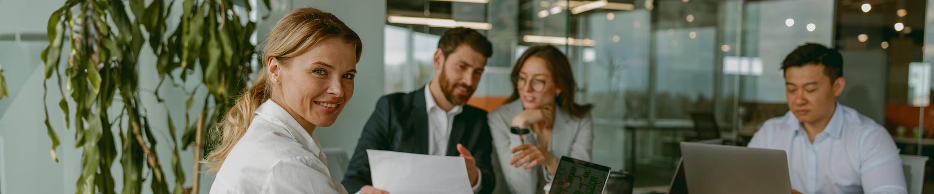 Drei Personen sitzen an einem Tisch in einem Büro, eine Person schaut zur Kamera und lächelt
