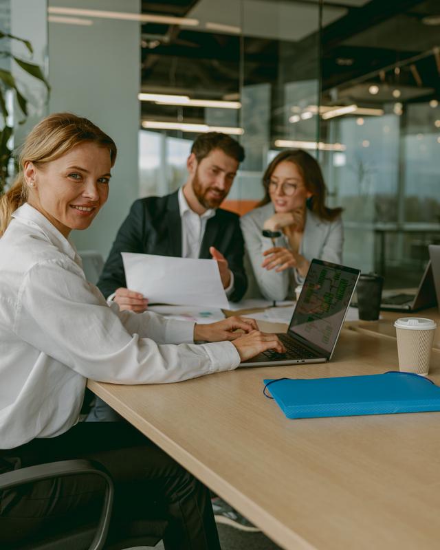 Drei Personen sitzen an einem Tisch in einem Büro, eine Person schaut zur Kamera und lächelt Drei Personen sitzen an einem Tisch in einem Büro, eine Person schaut zur Kamera und lächelt