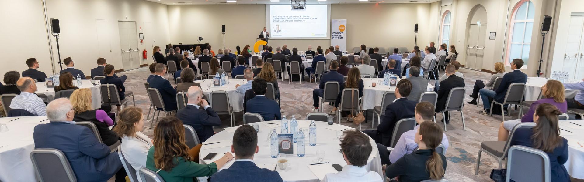 GRC_DAY_2025 Konferenzraum mit mehreren runden Tischen, an denen Teilnehmer sitzen und einem Vortragenden, der vor einer Leinwand präsentiert