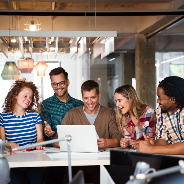 Sechs junge Erwachsene sitzen um einen Tisch und schauen gemeinsam lachend auf einen Laptop in einem modernen Büro