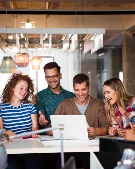Sechs junge Erwachsene sitzen um einen Tisch und schauen gemeinsam lachend auf einen Laptop in einem modernen Büro