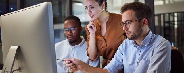 Drei Personen sitzen an einem Schreibtisch und schauen gemeinsam auf einen großen Computerbildschirm Drei Personen sitzen an einem Schreibtisch und schauen gemeinsam auf einen großen Computerbildschirm