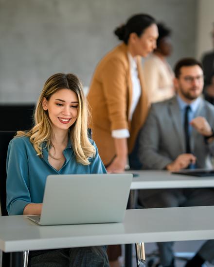 Junge Frau in blauem Hemd arbeitet lächelnd an Laptop in modernem Büro mit Kollegen im Hintergrund