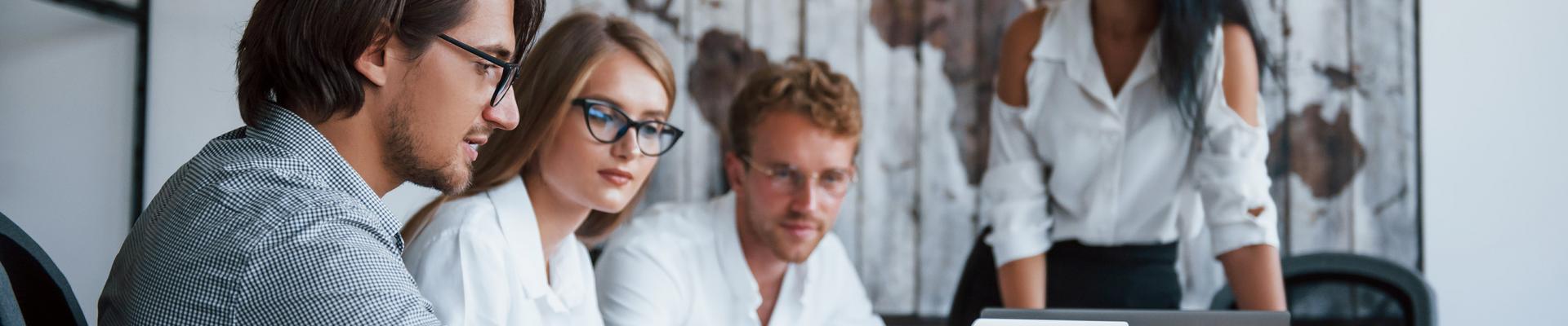 Drei Personen sitzen an einem Tisch und schauen auf einen Laptop, eine vierte Person steht im Hintergrund vor einer Wand mit einem großen Bild Drei Personen sitzen an einem Tisch und schauen auf einen Laptop, eine vierte Person steht im Hintergrund vor einer Wand mit einem großen Bild