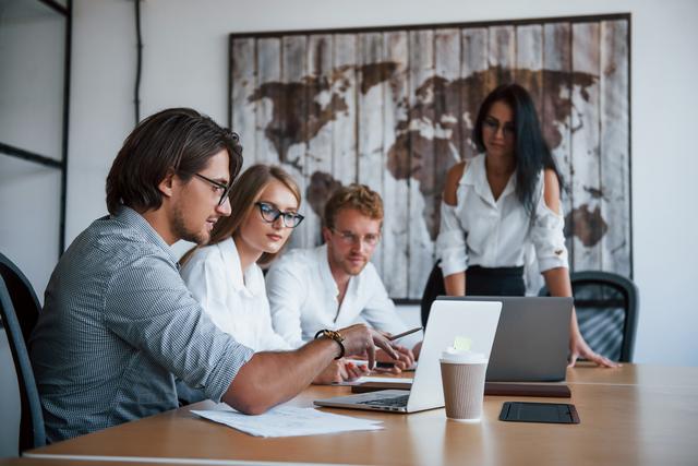 Drei Personen sitzen an einem Tisch und schauen auf einen Laptop, eine vierte Person steht im Hintergrund vor einer Wand mit einem großen Bild