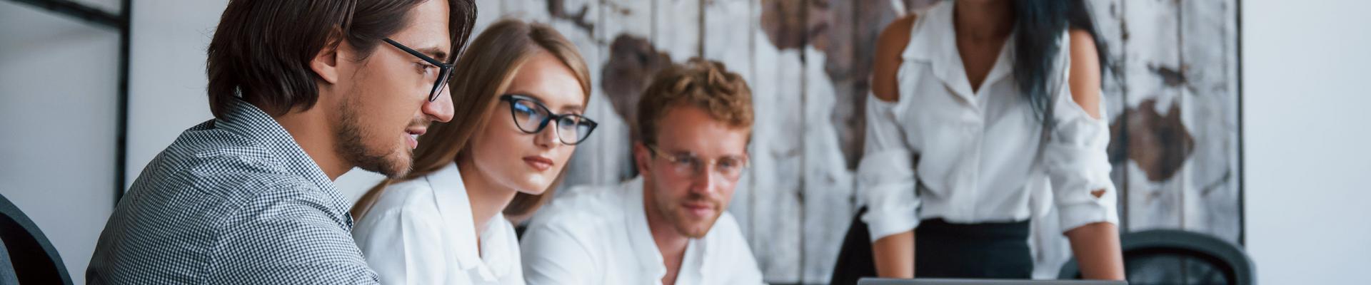 Drei Personen sitzen an einem Tisch und schauen auf einen Laptop, eine vierte Person steht im Hintergrund vor einer Wand mit einem großen Bild Drei Personen sitzen an einem Tisch und schauen auf einen Laptop, eine vierte Person steht im Hintergrund vor einer Wand mit einem großen Bild